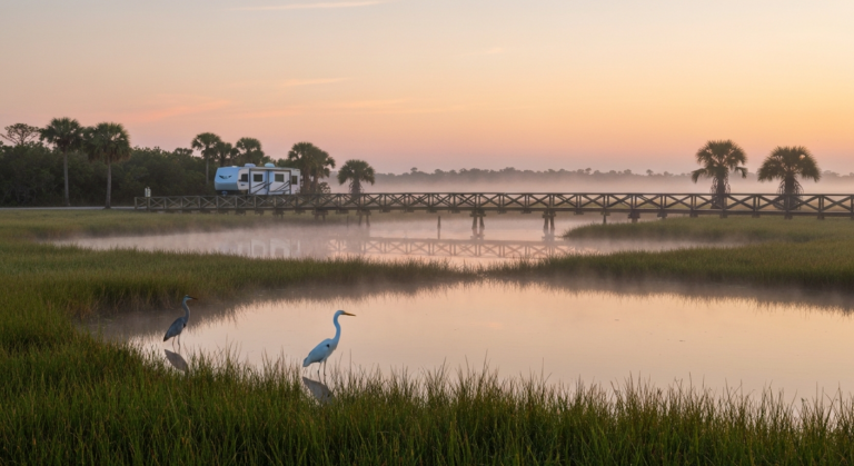 Sunrise over the sawgrass marsh and cypress boardwalk at Loxahatchee National Wildlife Refuge with wading birds and a travel-trailer RV at a distant trailhead